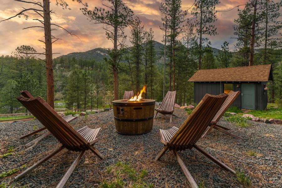 Barrel fire pit ringed by wooden lounge chairs among tall pines at sunset