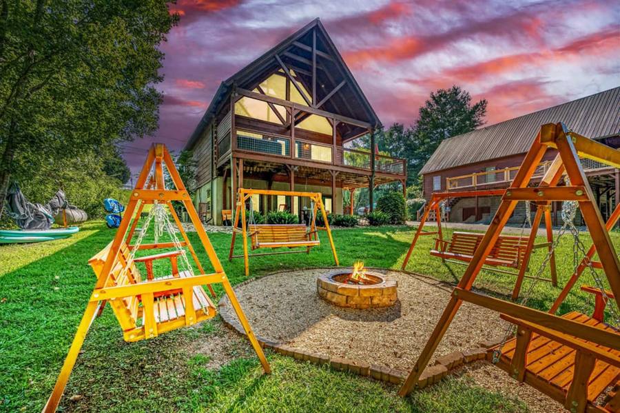A-frame cabin at sunset with a fire pit circled by wooden porch swings